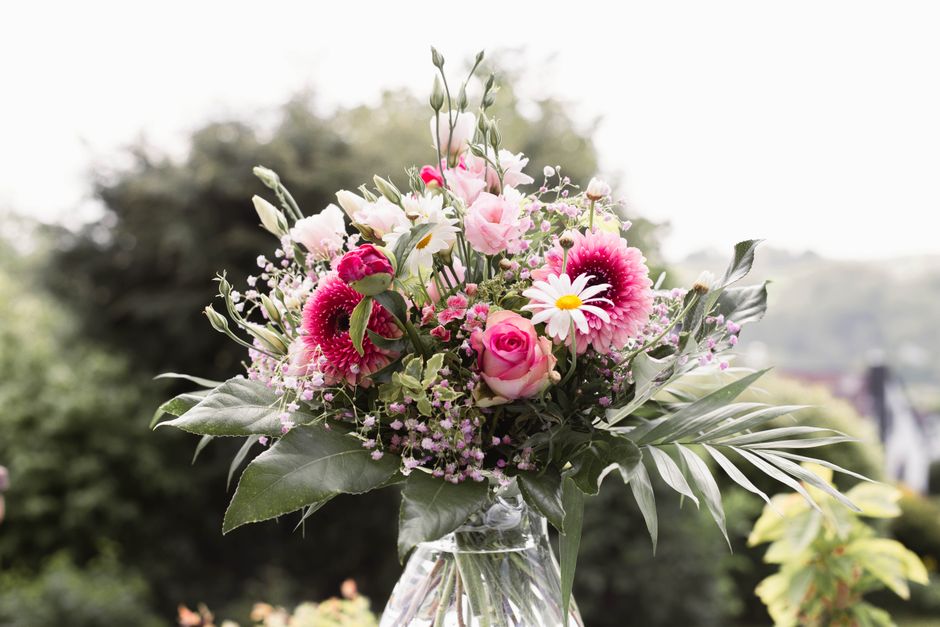 A vibrant floral arrangement featuring pink roses, white daisies with yellow centers, and pink gerbera daisies, all arranged in a clear glass vase. The bouquet is accented with green foliage, includin