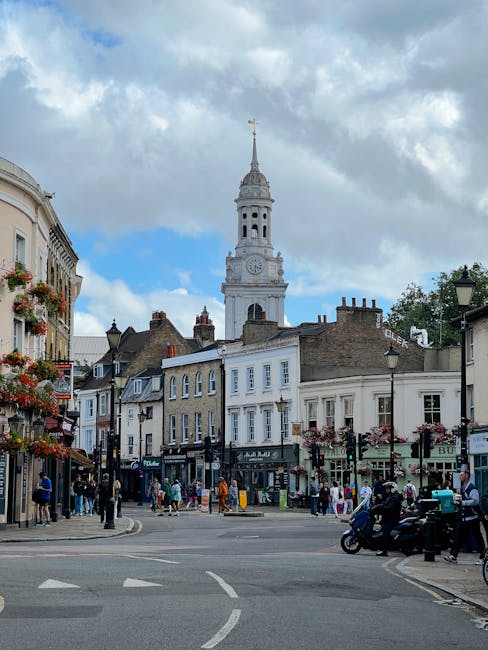 This image depicts a vibrant street scene in a historic town centre, featuring a large clock tower with a spire in the background under a partly cloudy sky. The foreground shows a busy intersection wi