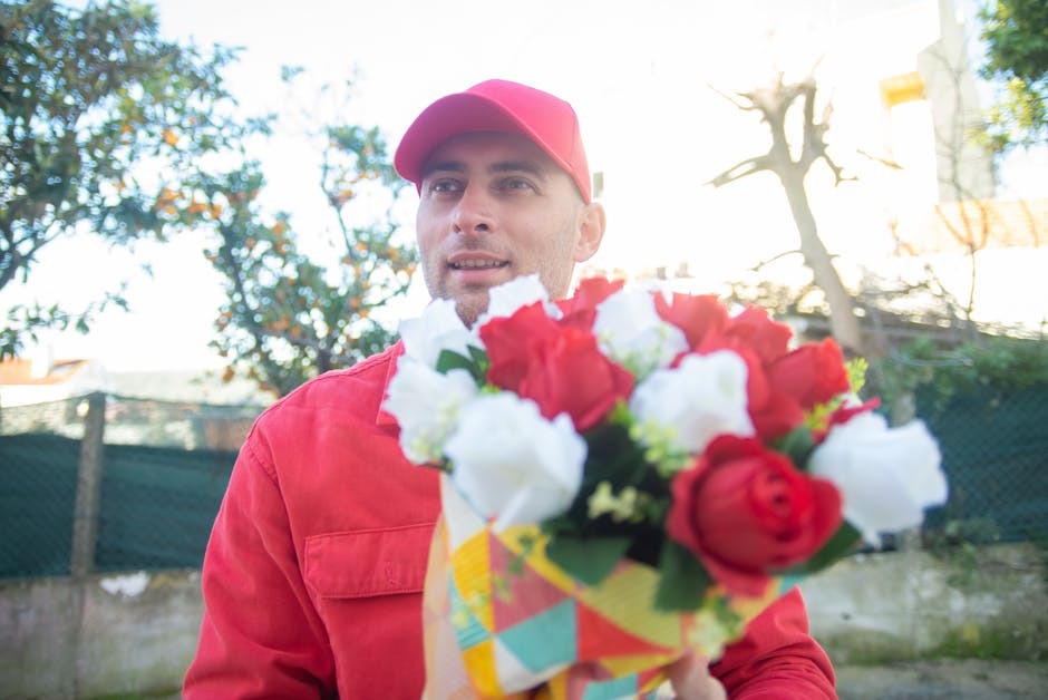 A man in a red jacket and matching red cap holds a vibrant floral bouquet close to the camera during daytime. The bouquet features an assortment of fresh roses and lisianthus in red and white, arrange