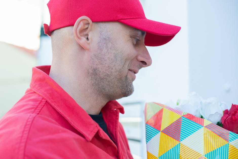 A man in a red cap and matching red jacket is holding a colorful geometric-patterned gift bag filled with white and pink flowers. The flowers include roses with ruffled petals and vibrant hues. The im