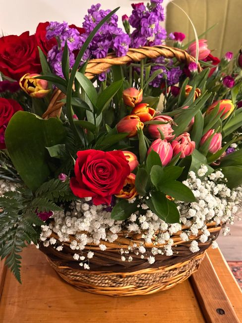 A woven wicker basket filled with a vibrant floral arrangement including a single large red rose, pink tulips, and purple stock flowers, accented by white gypsophila and lush green foliage. The flower