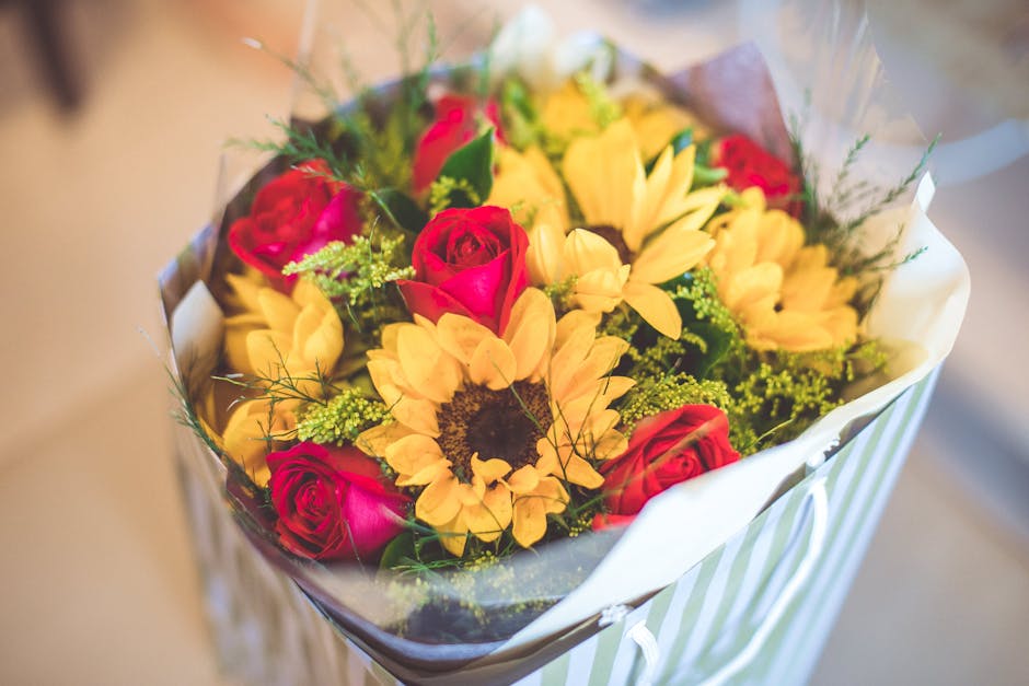 A bouquet of fresh flowers arranged with a combination of vibrant pink roses, creamy yellow sunflowers, and delicate green foliage, wrapped in clear cellophane and white paper with subtle green stripe
